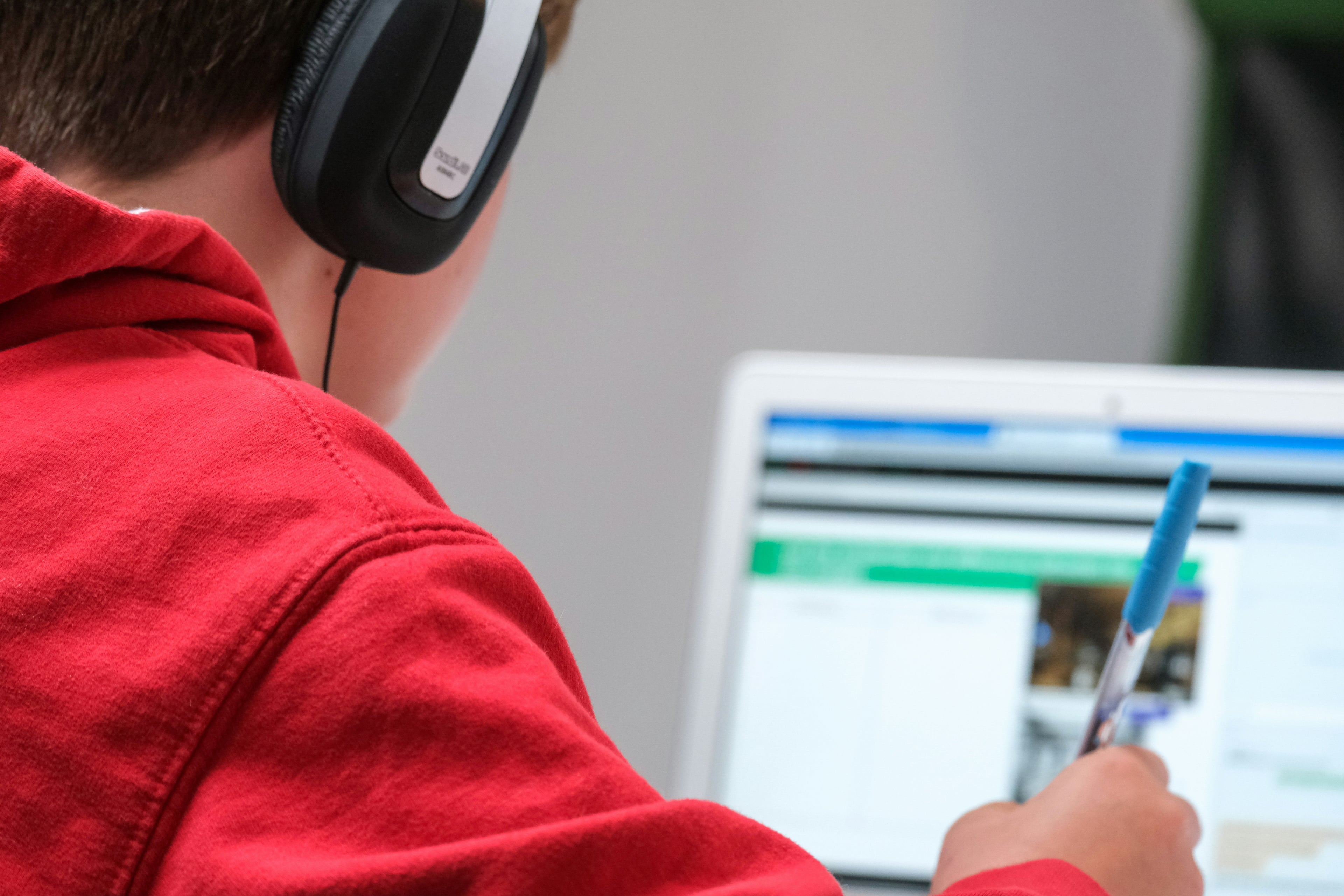 Student with headphones and red hoodie looking at laptop, holding a pen.